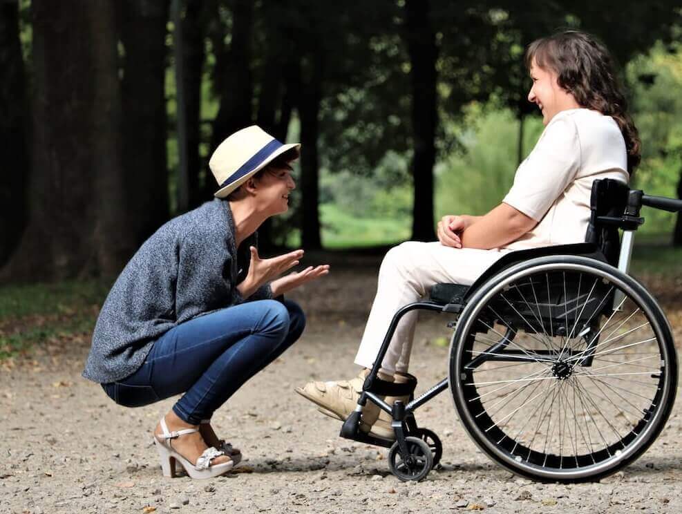 Two women smiling, with one kneeling and talking to the other, who is sitting in a wheelchair in a park setting. - Home Instead