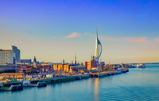 A waterfront cityscape with the Spinnaker Tower and surrounding buildings at sunset, calm water in the foreground. - Home Instead