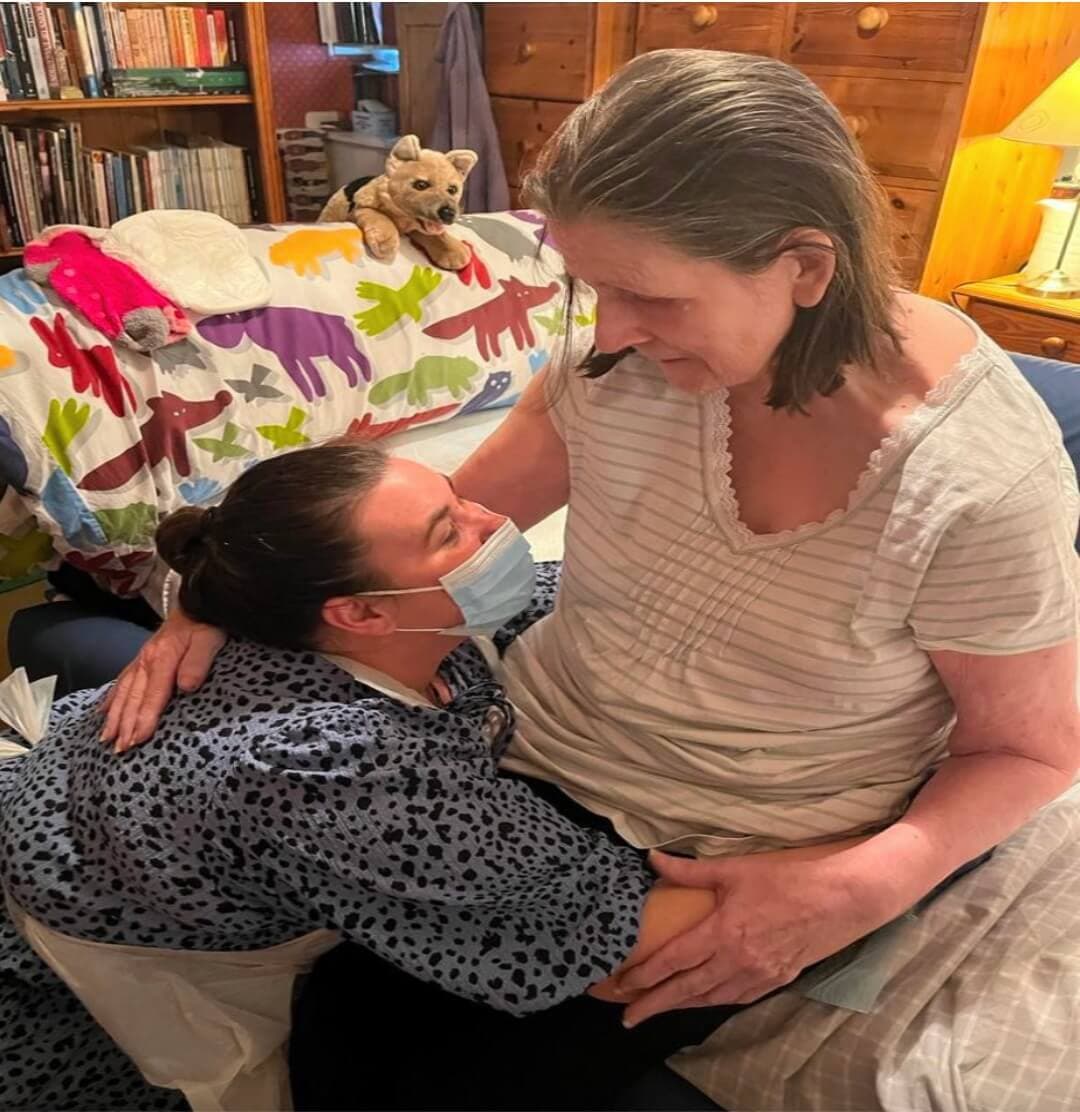A caregiver wearing a mask embraces an elderly woman sitting on a bed covered with a colorful dinosaur blanket. - Home Instead