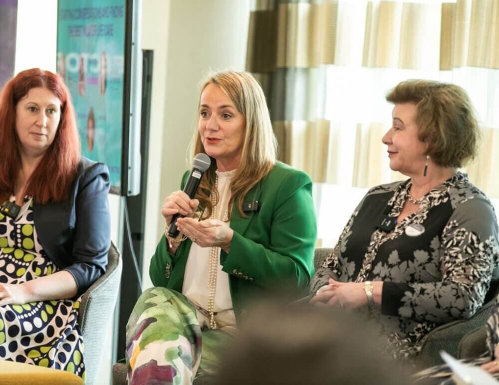 Three women are seated; one in a green blazer is speaking into a microphone, with others listening attentively. - Home Instead