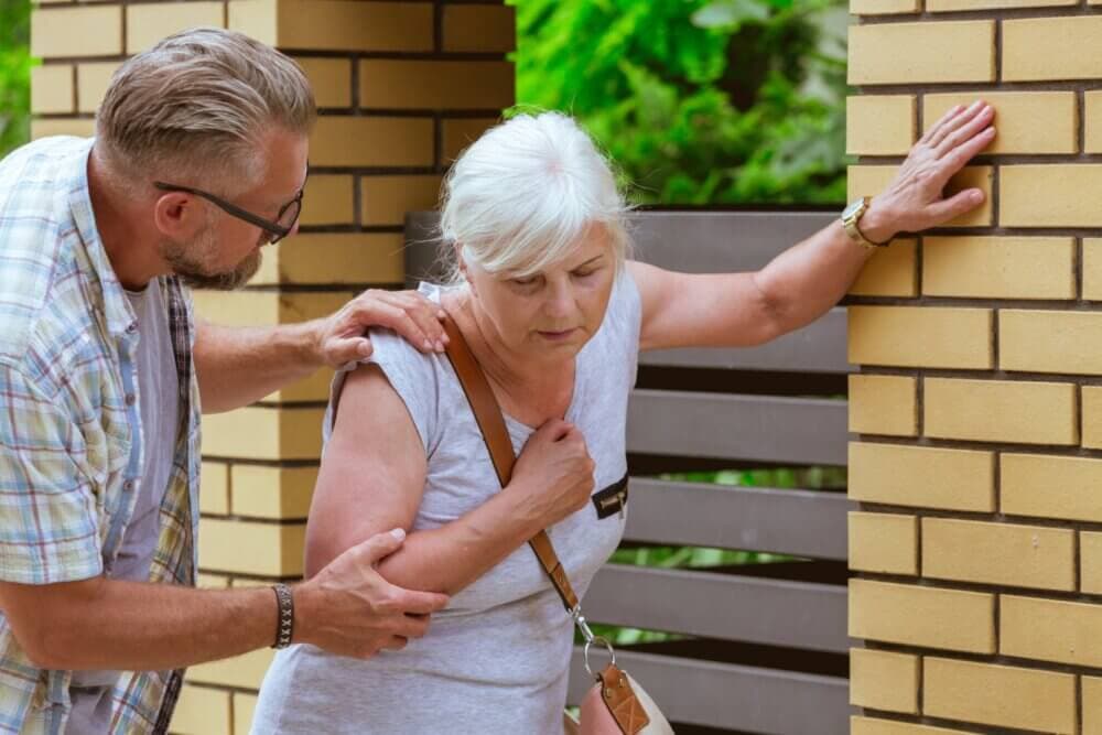 A man assists an elderly woman who appears to be in distress, leaning against a brick wall. - Home Instead