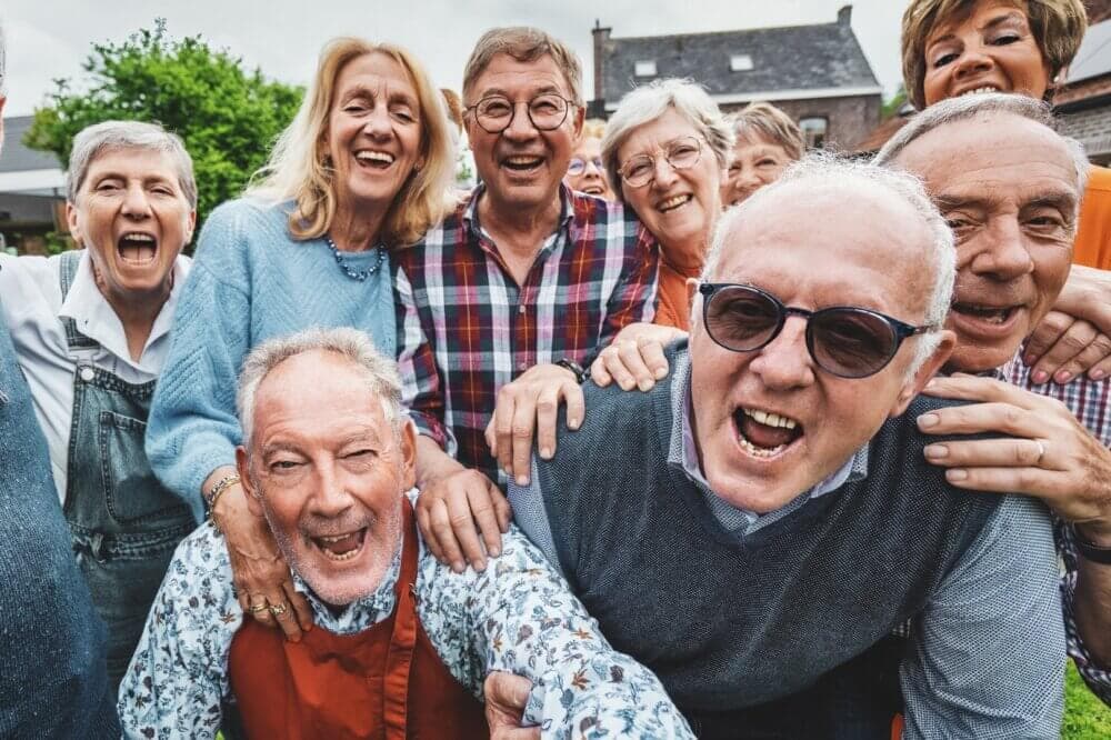 A group of elderly people, smiling and having fun outdoors, some wearing glasses, casual clothes, and bright expressions. - Home Instead