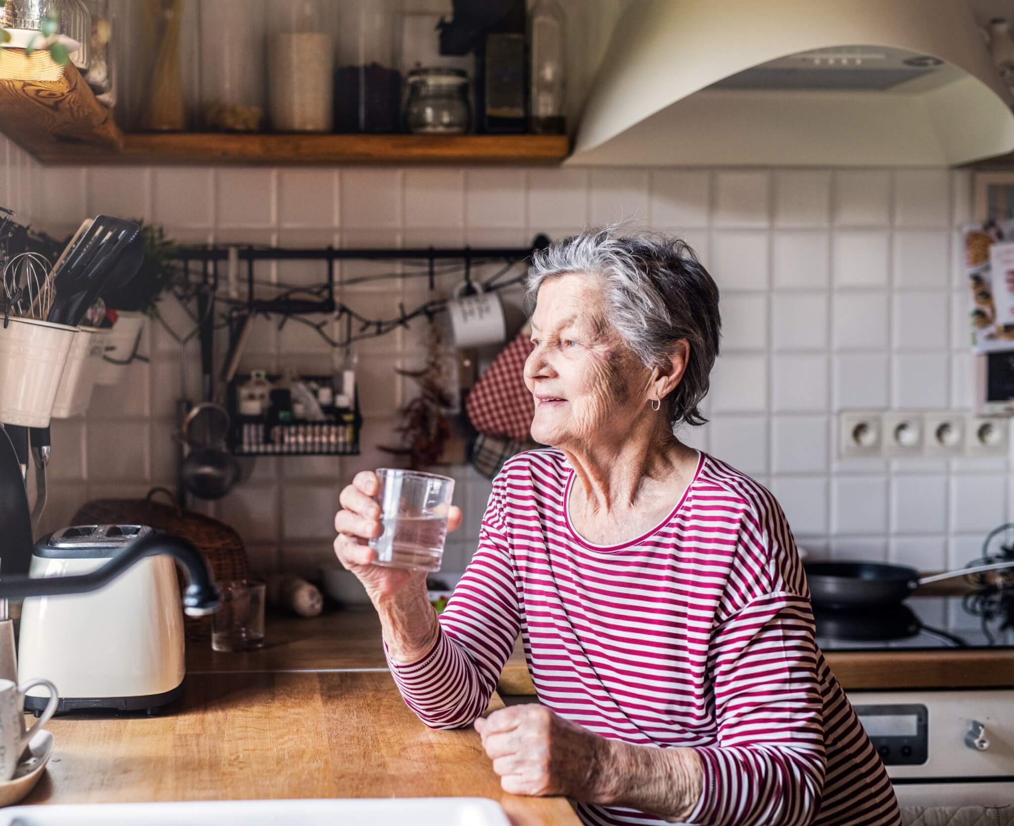 An elderly woman in a red-striped shirt holds a glass of water while sitting in a kitchen with white tile walls. - Home Instead