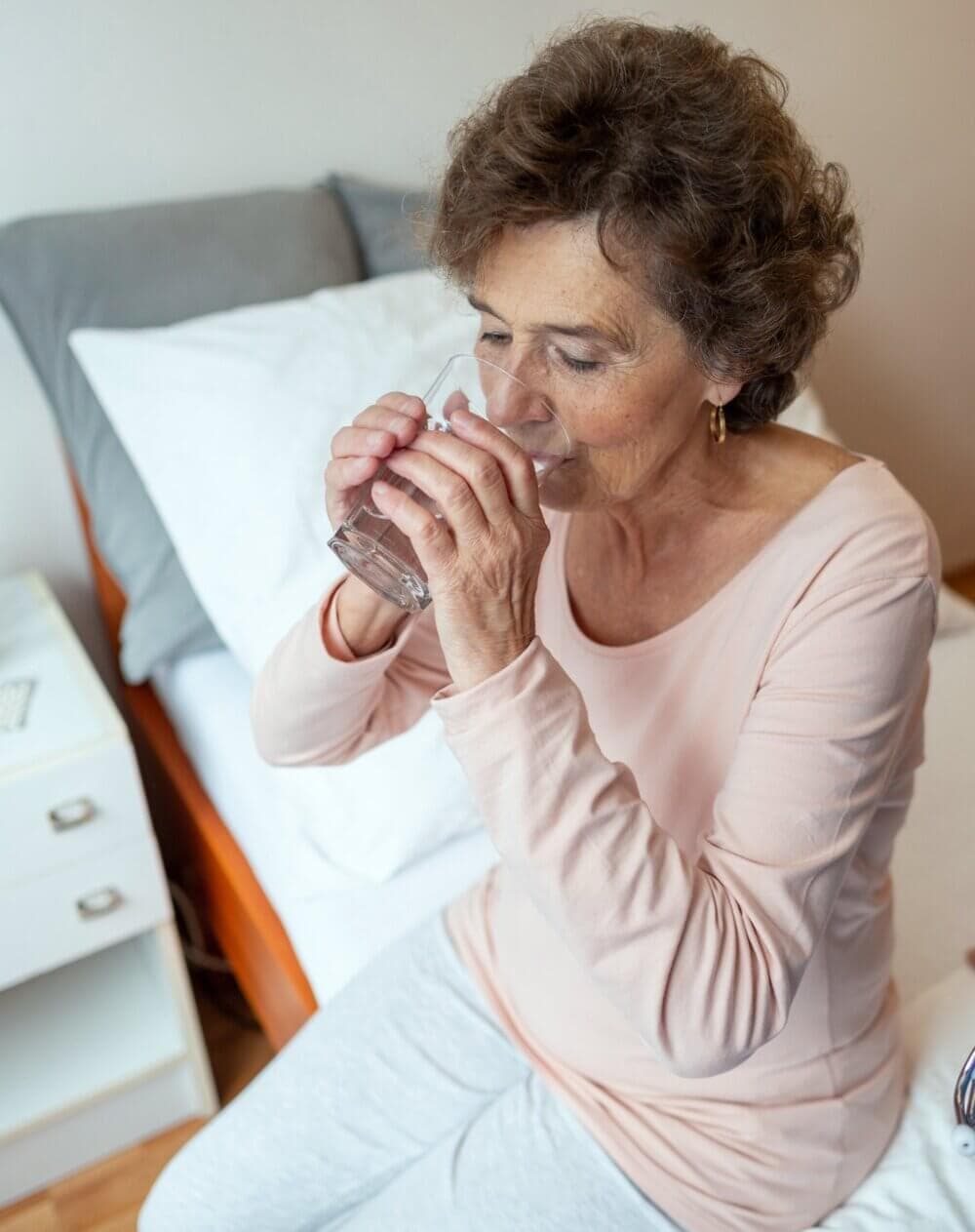 Older woman sitting on a bed, drinking water from a glass. - Home Instead