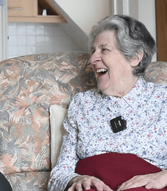 An elderly woman with gray hair, wearing a floral blouse, smiles while sitting on a patterned sofa. - Home Instead