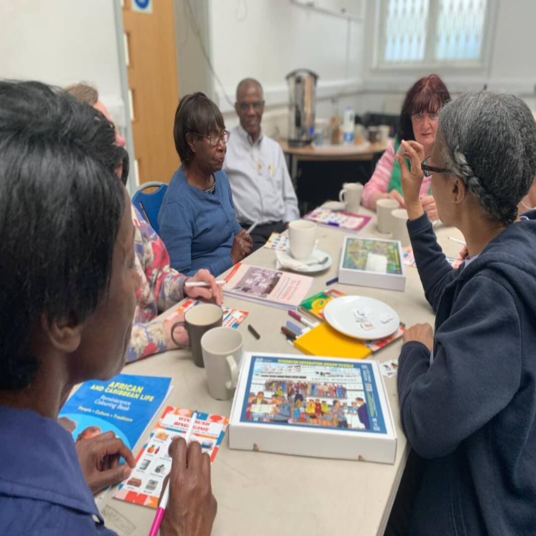 A group of people sitting around a table, engaging in conversation and looking at books and games. - Home Instead