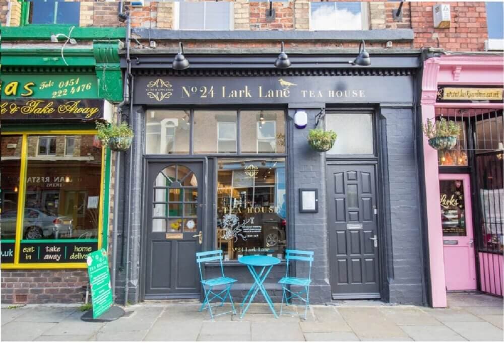Front of a tea house with black facade, blue chairs and table outside, between two colorful storefronts. - Home Instead