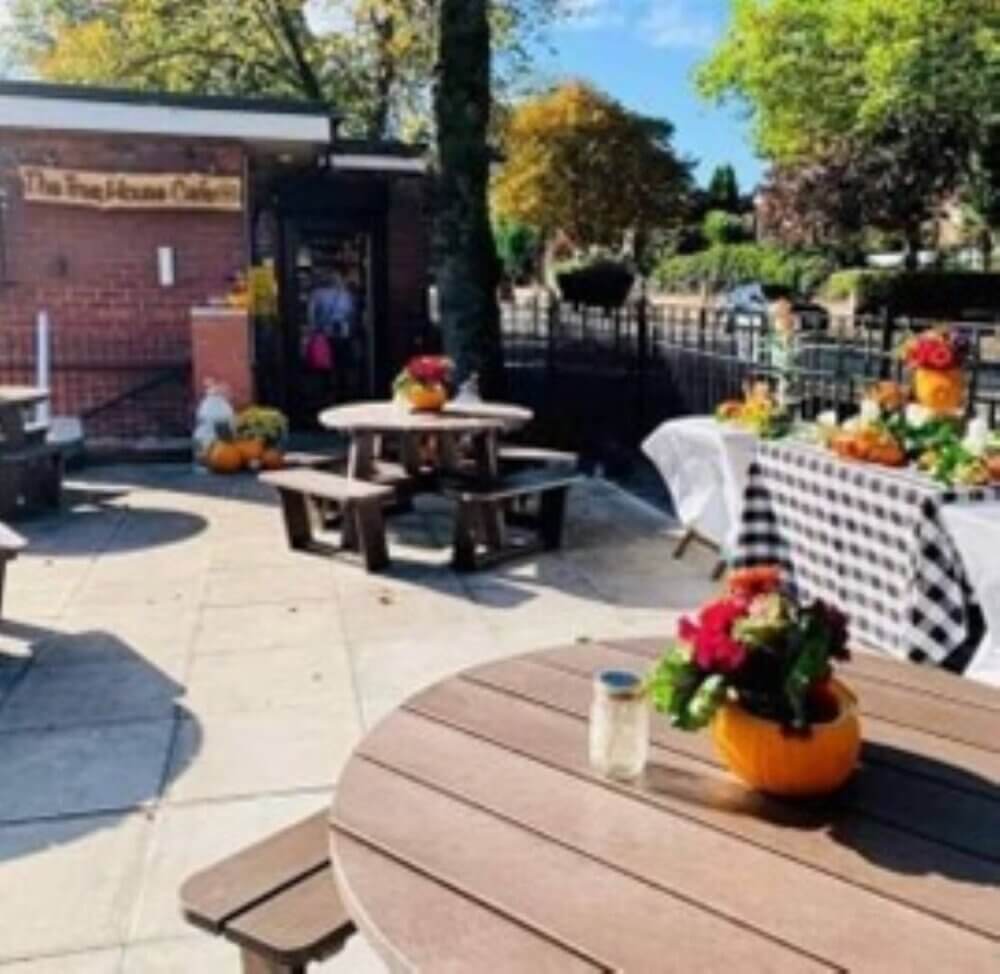 Outdoor cafe with picnic tables, flower arrangements in pumpkins, checkered tablecloths, and a backdrop of trees and shrubs. - Home Instead