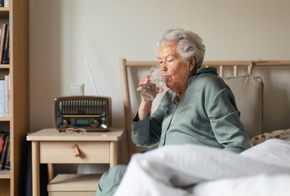 Older woman in green pajamas sits on a bed and drinks water. A vintage radio sits on the bedside table. - Home Instead