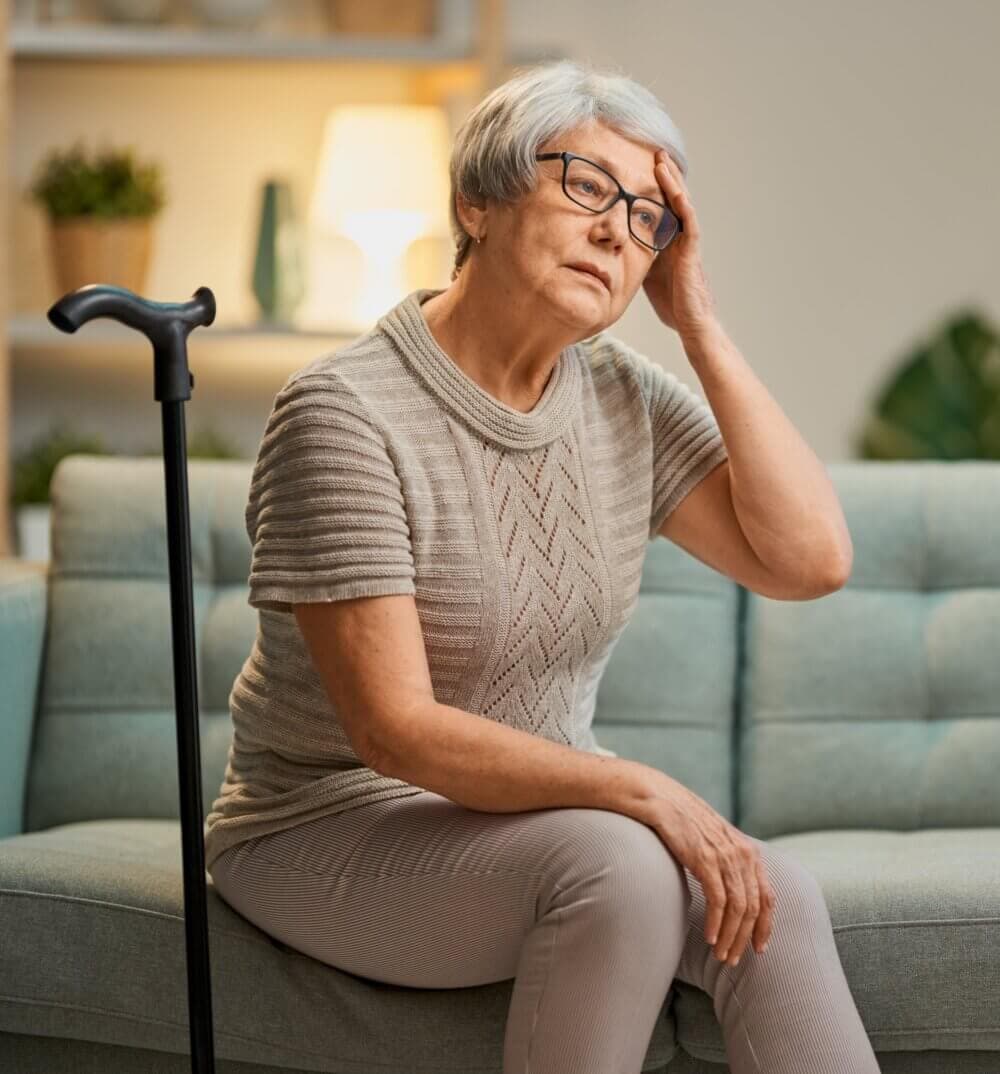 Elderly woman with glasses and a cane sitting on a couch, looking pensive with her hand on her forehead. - Home Instead
