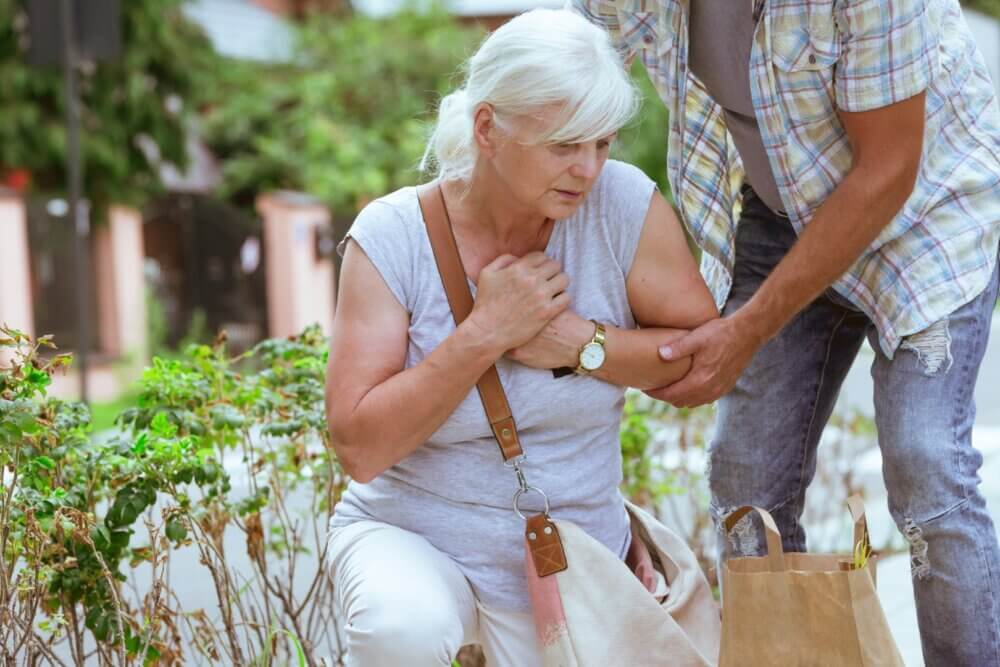 An elderly woman appears to be in distress and is being helped by a man. They are outdoors with bags around them. - Home Instead