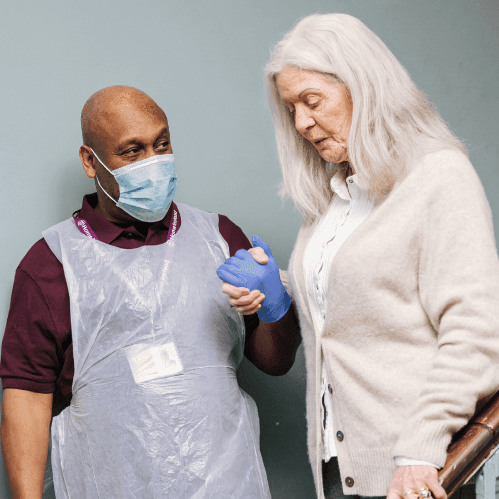 Healthcare worker in PPE assisting an elderly woman with white hair as she holds a cane. - Home Instead