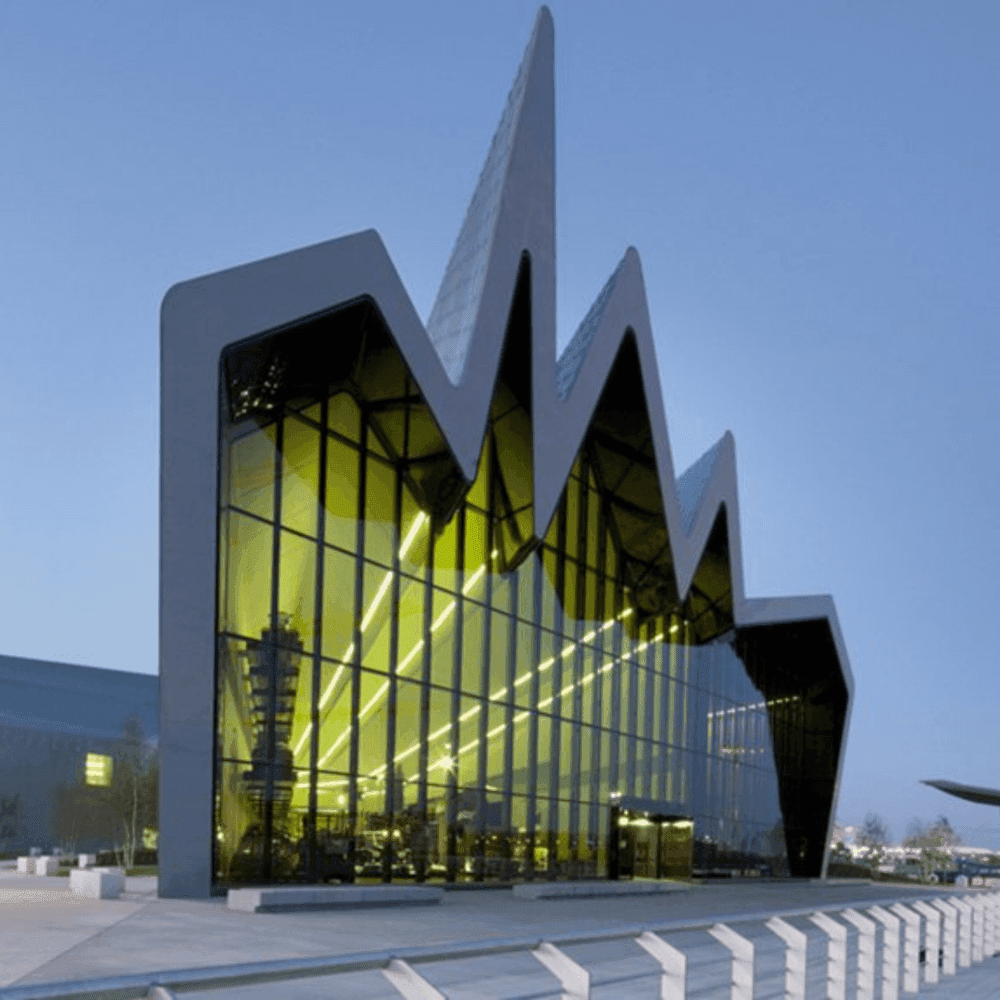 Modern building with a unique, jagged roof design and large glass facade illuminated by yellow lights at dusk. - Home Instead