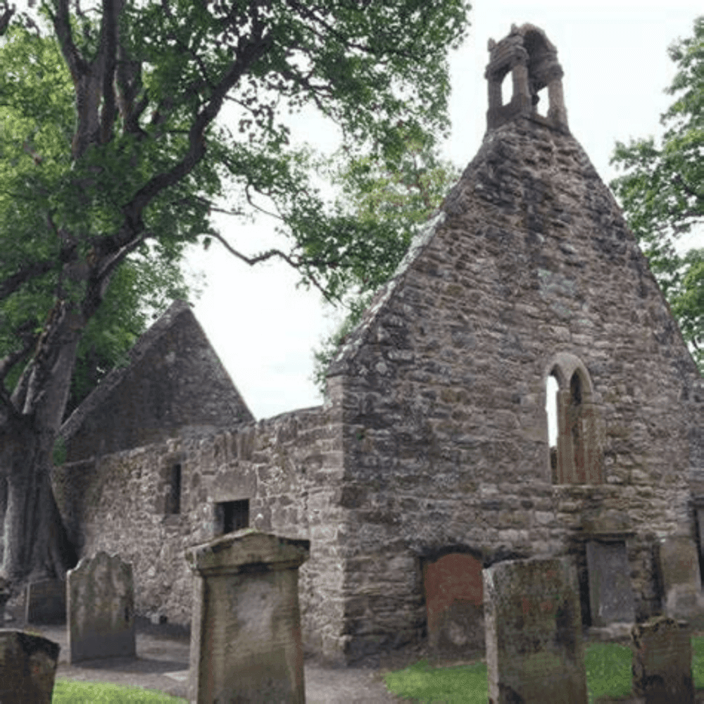 Ancient stone building with trees around, featuring old headstones in the foreground and a bell gable at the top. - Home Instead