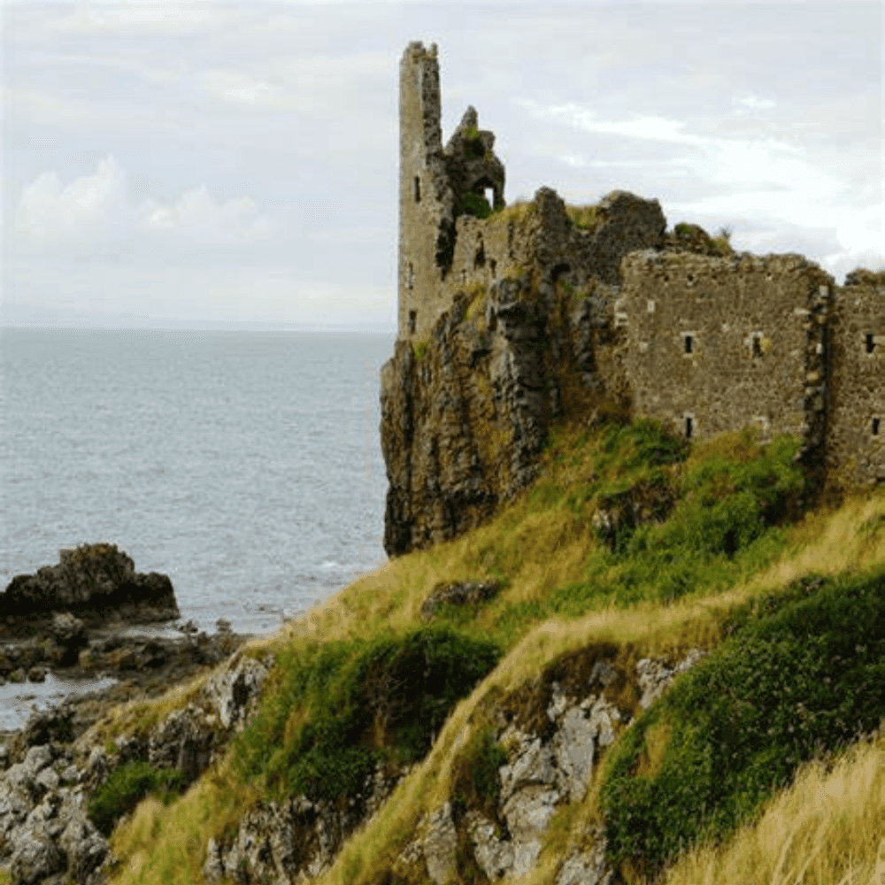 A ruined stone castle stands on a rocky cliff by the sea with grassy slopes in the foreground under a cloudy sky. - Home Instead