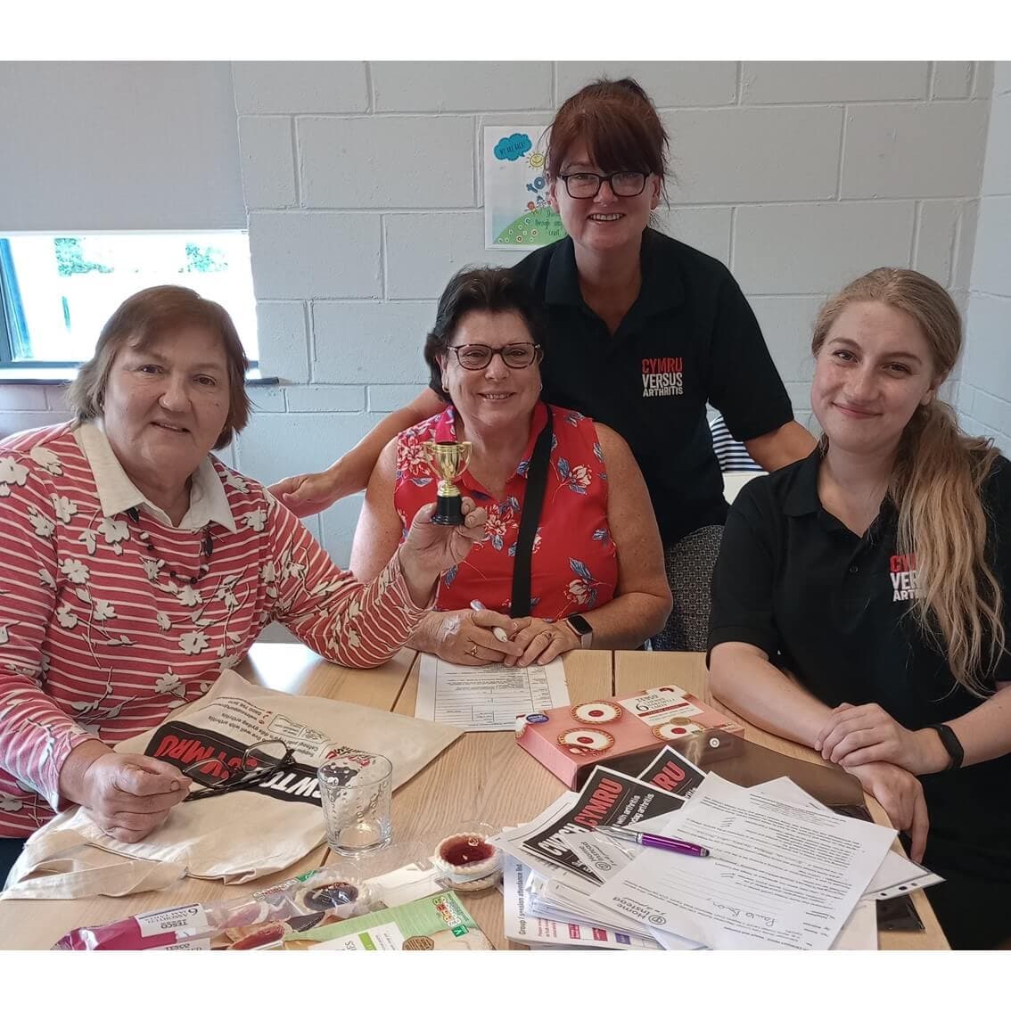 Four women smile for the camera while sitting at a table filled with papers and crafting materials. - Home Instead