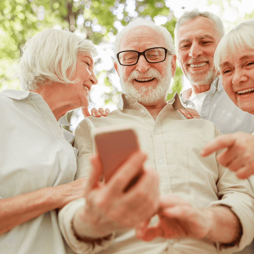 Four elderly people smiling and looking at a smartphone outdoors with greenery in the background. - Home Instead