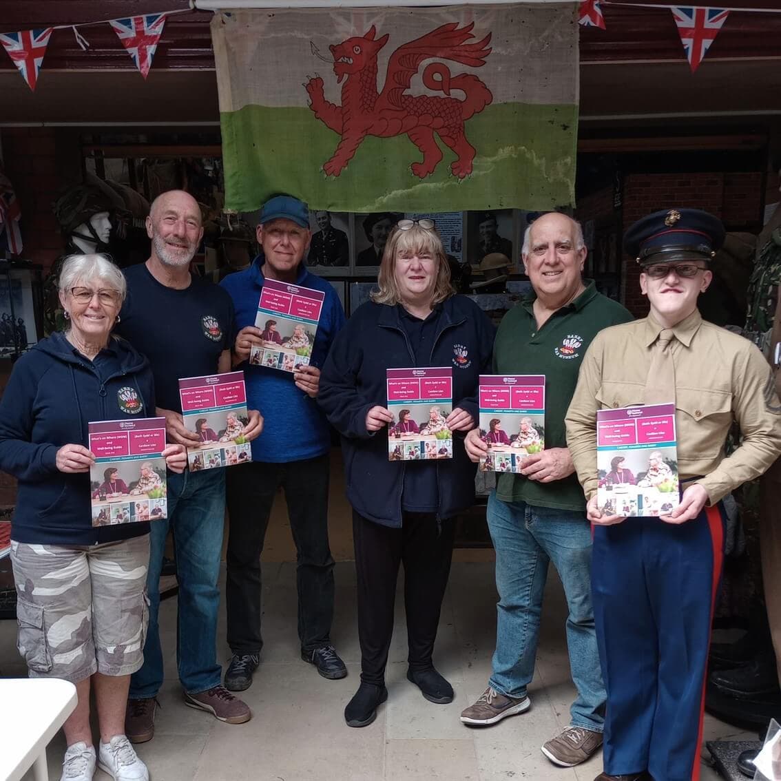Six people holding community newsletters, standing in front of a wall with a Welsh flag and some British flags. - Home Instead