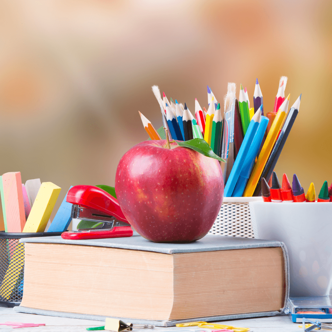 An apple, colorful pencils, and a red stapler on a book with various office supplies in the background. - Home Instead