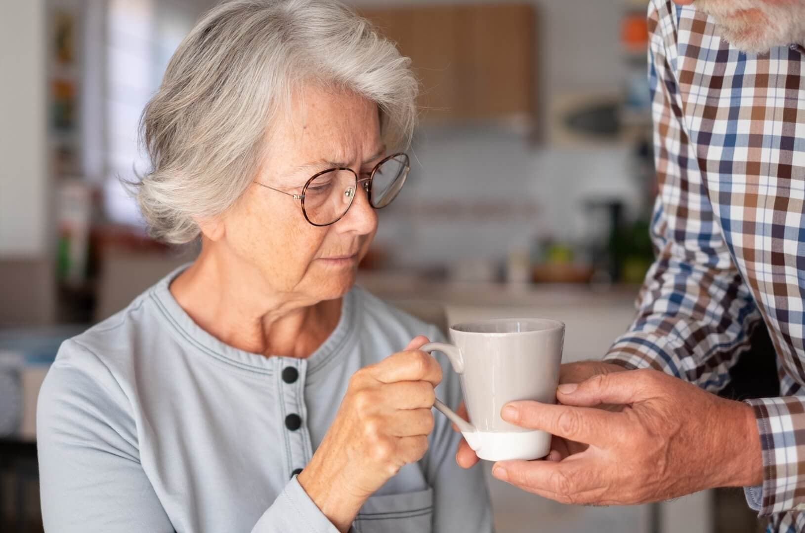 Elderly woman with gray hair and glasses holding a coffee mug, assisted by another person in a checkered shirt. - Home Instead