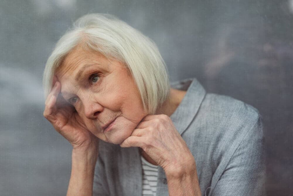 Elderly woman with short white hair leaning against a glass pane, looking contemplative, wearing a light gray top. - Home Instead