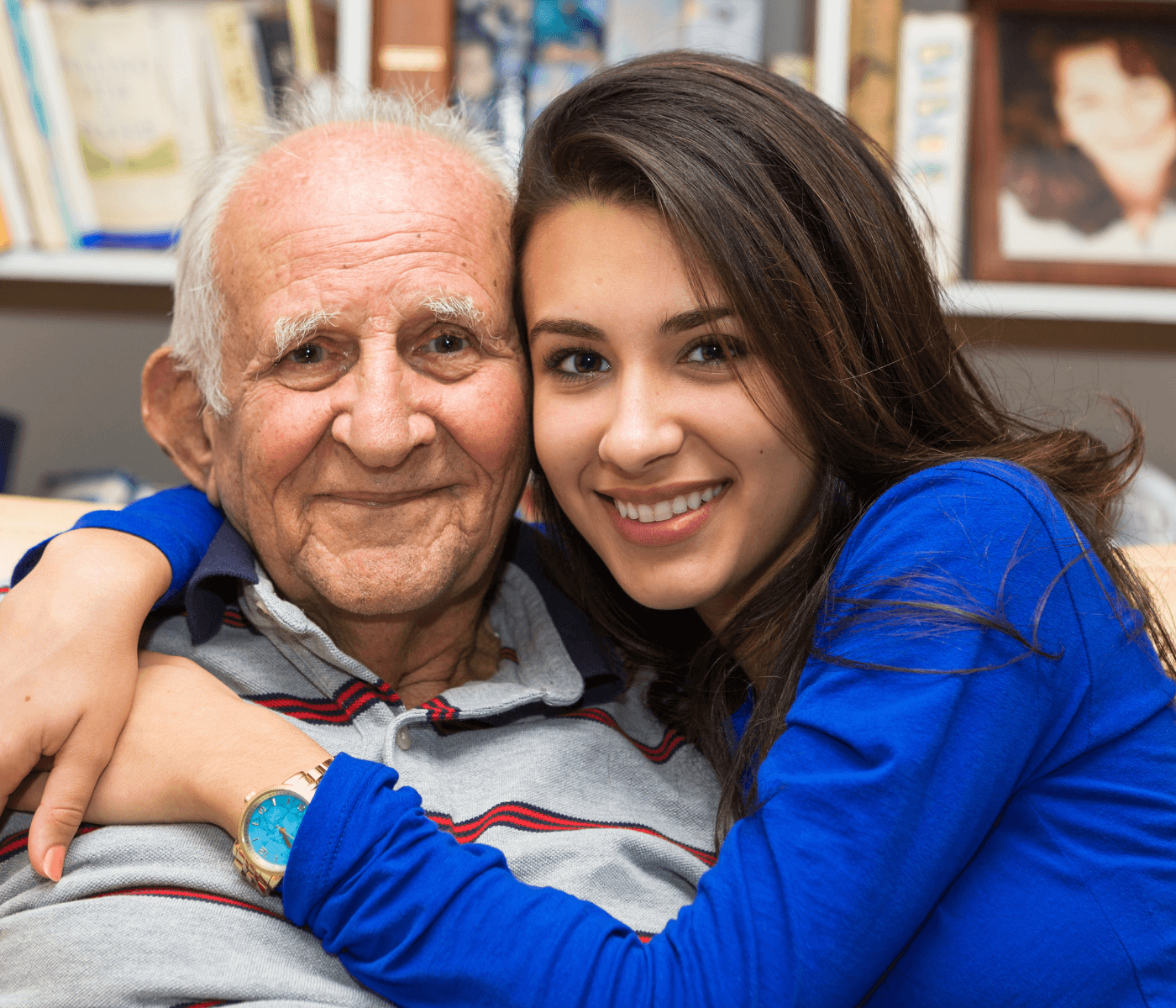 A smiling young woman hugging an elderly man, both wearing blue shirts, with bookshelves in the background. - Home Instead