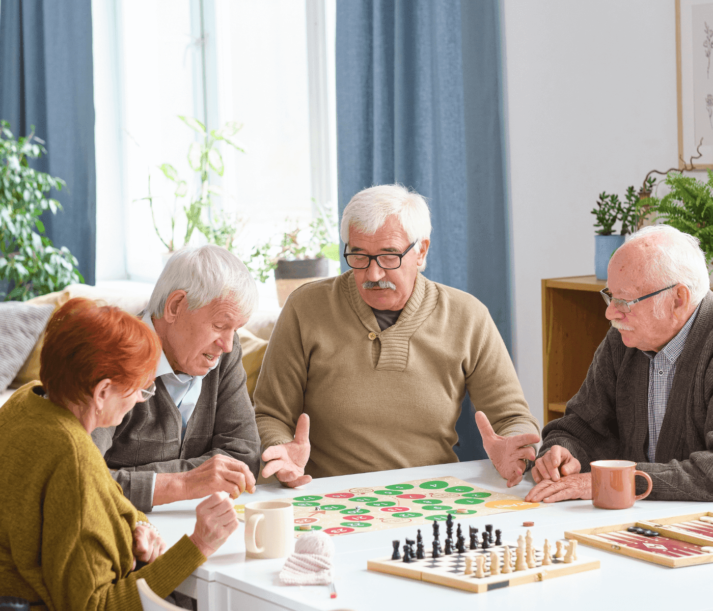 A group of elderly people playing a board game and talking at a table, with a chessboard and coffee mugs near them. - Home Instead