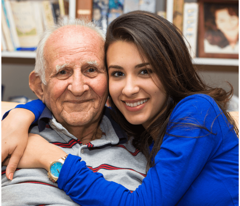An elderly man and a young woman smile for the camera as they hug, with bookshelves and framed photos in the background. - Home Instead