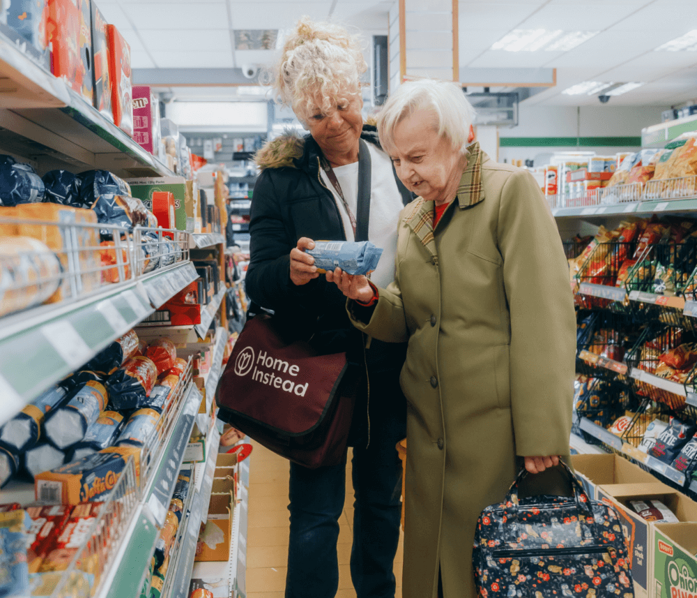 A caregiver assists an elderly woman in choosing an item at a grocery store aisle. - Home Instead