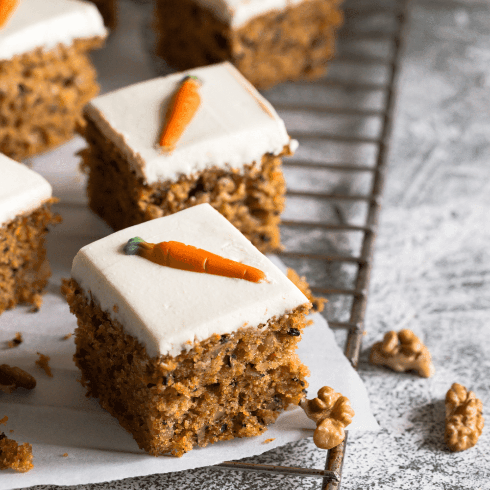 Carrot cake squares topped with cream cheese frosting and fondant carrot decorations on a cooling rack. - Home Instead