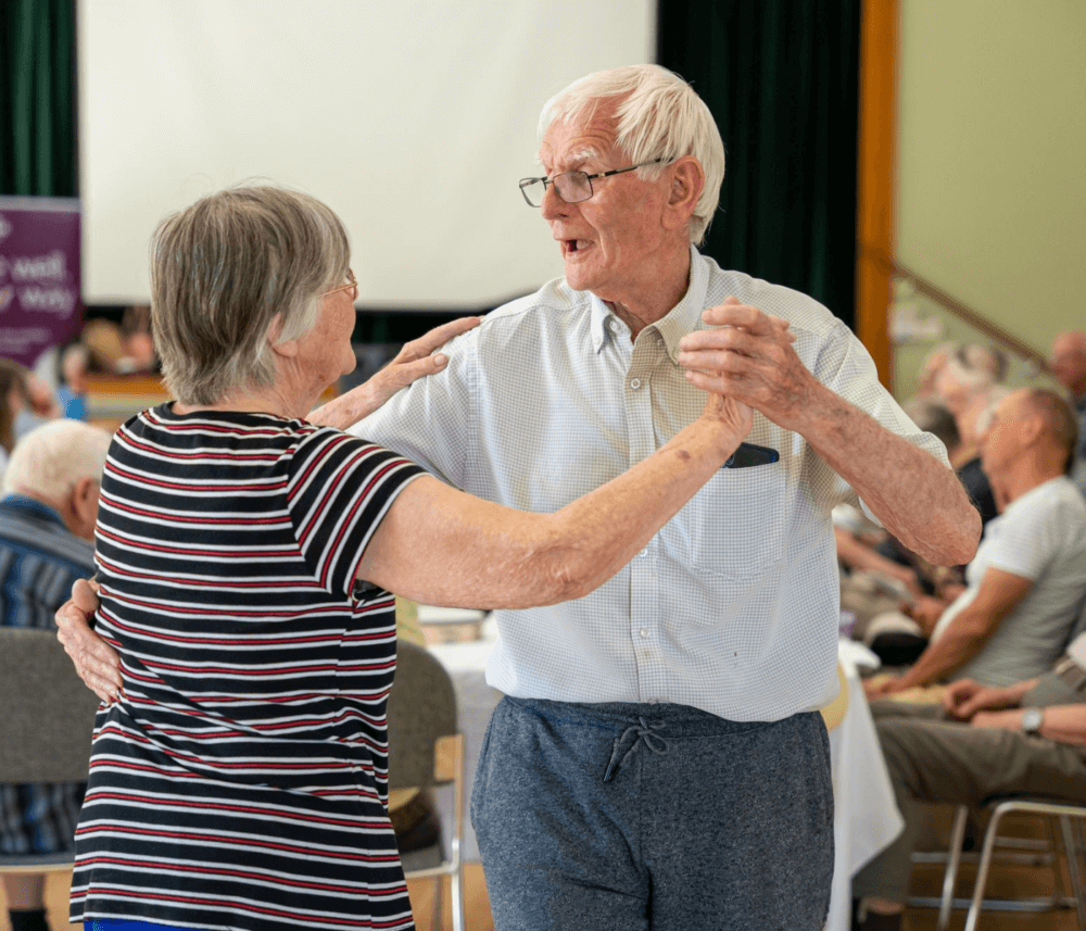 Elderly couple dances together in a community hall, smiling and holding hands, surrounded by other seated seniors. - Home Instead