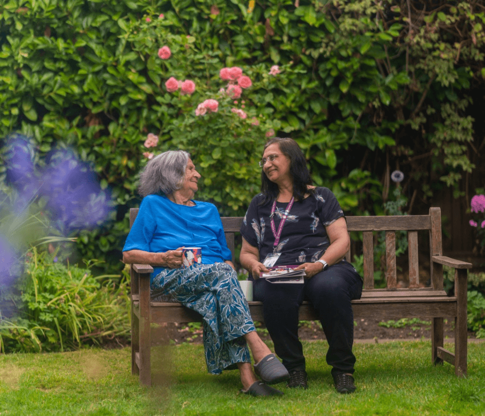Two women sitting on a bench in a garden, smiling and chatting, surrounded by green foliage and blooming flowers. - Home Instead