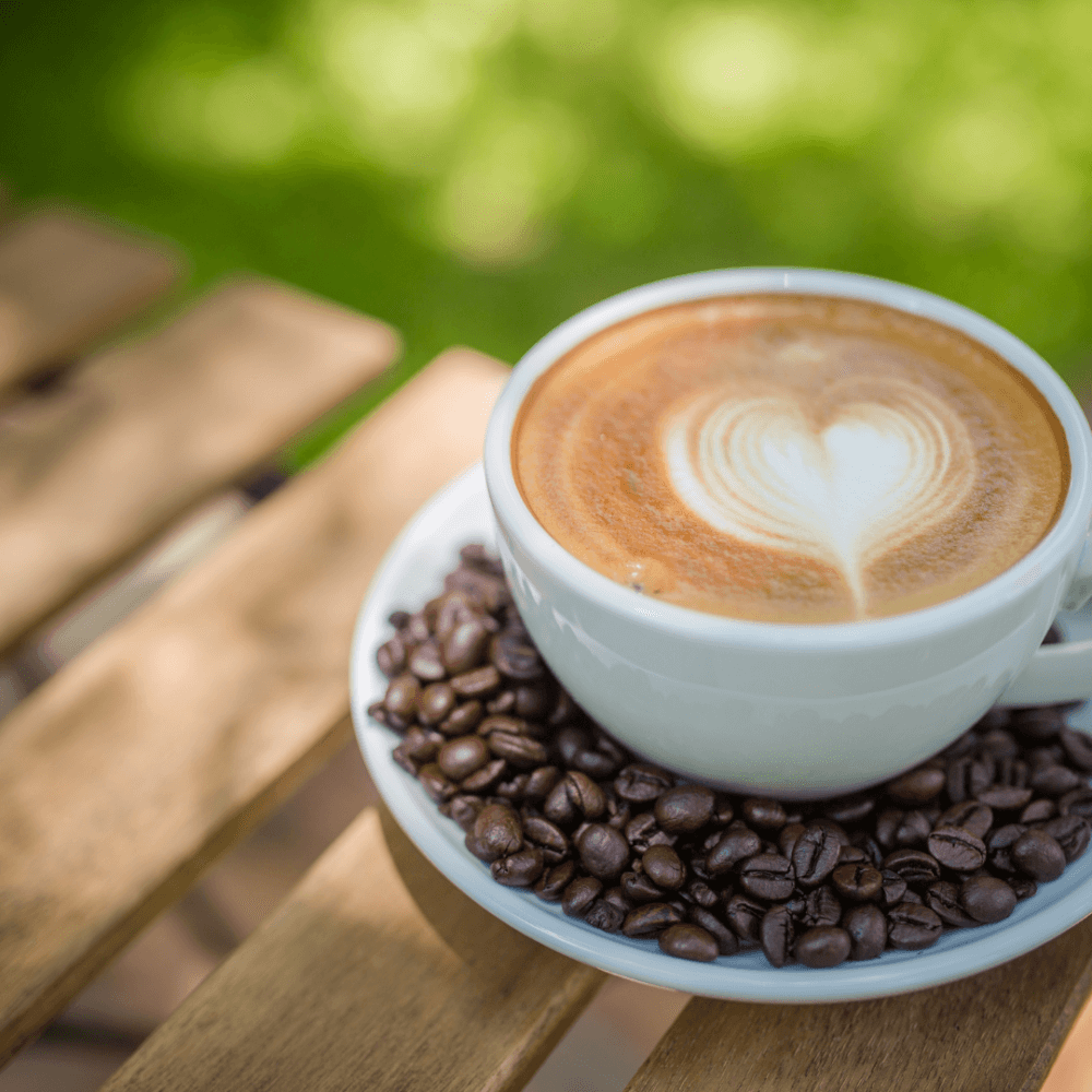 A cup of coffee with heart-shaped latte art, placed on a saucer filled with coffee beans, resting on a wooden table. - Home Instead