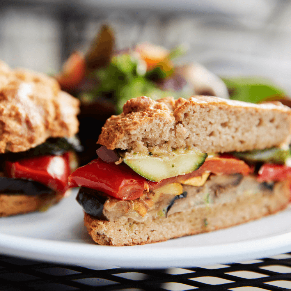 Close-up of a vegetable sandwich on a white plate with a salad in the background. - Home Instead