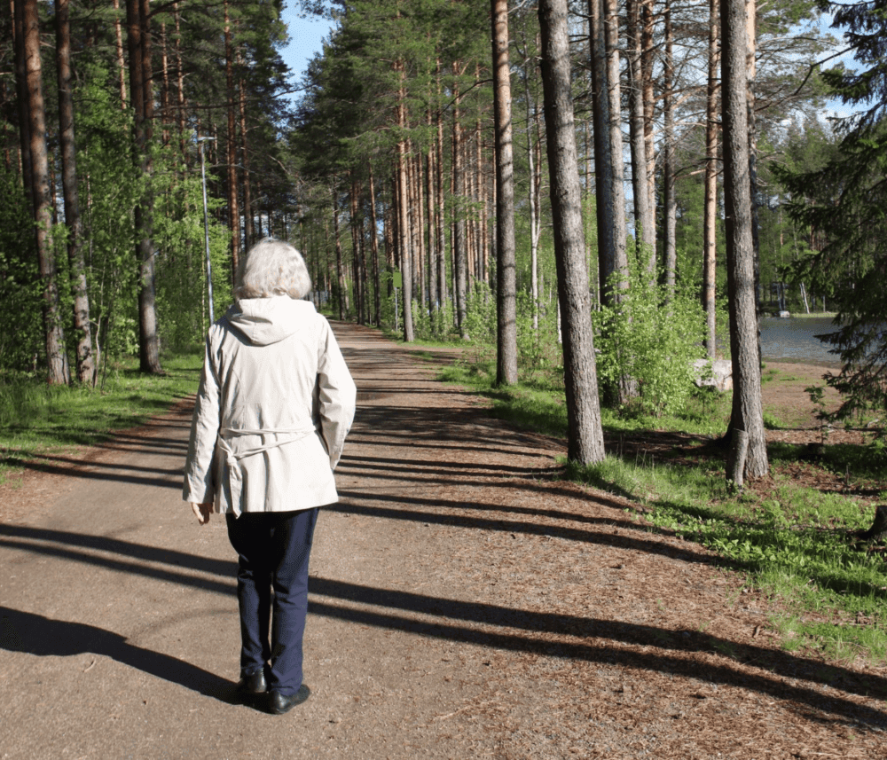A person with gray hair, in a white jacket, walks on a path through a forest with tall trees beside a body of water. - Home Instead