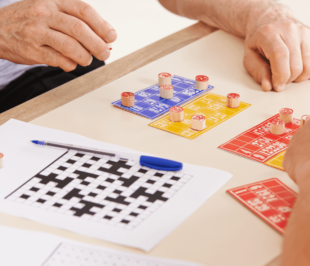 Two people playing a board game with numbered cards and wooden tokens next to a partially completed crossword puzzle. - Home Instead