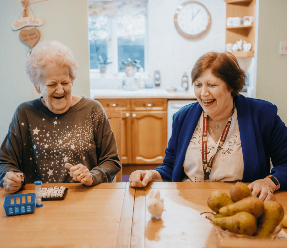Two women sitting at a table, smiling and playing a game in a cozy kitchen with pears and a toy on the table. - Home Instead