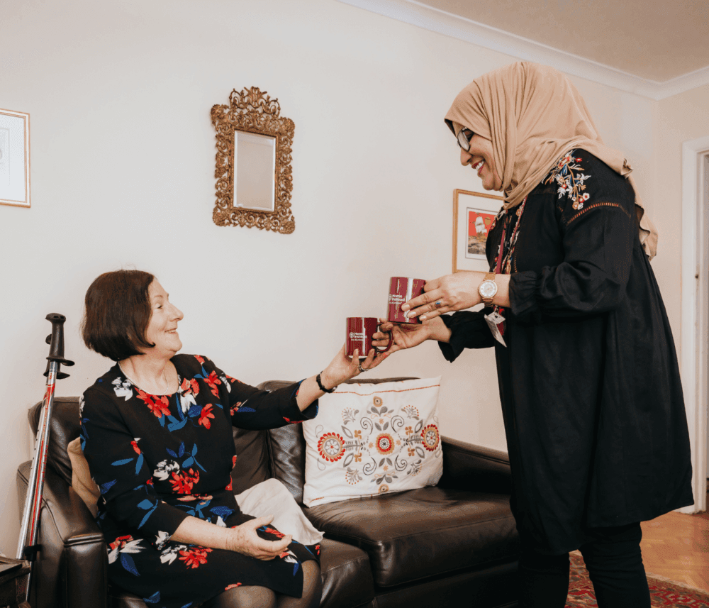 A woman in a hijab hands a drink to a seated woman with a walking stick, both smiling in a cozy living room. - Home Instead