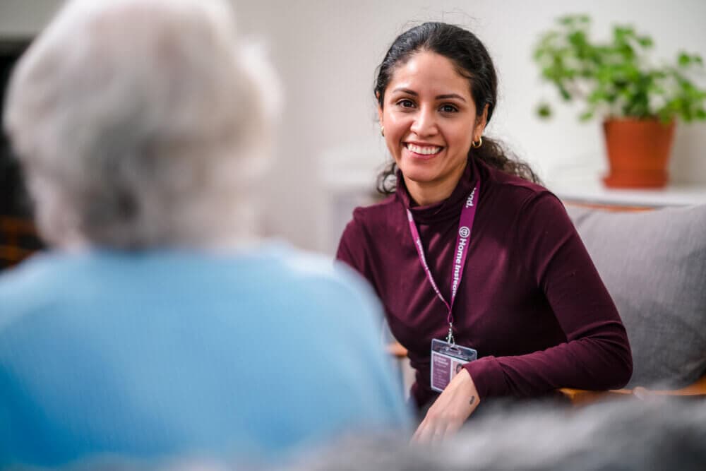 A smiling woman with a lanyard talks to an older person in a blue shirt, with a potted plant in the background. - Home Instead