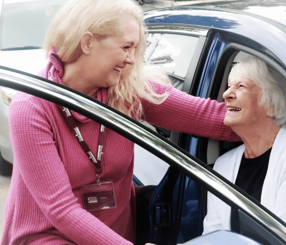 A smiling woman helps an elderly woman out of a car, both appear happy and engaged in conversation. - Home Instead
