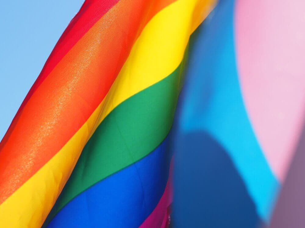 Close-up of a rainbow-colored Pride flag and a Transgender Pride flag waving against a clear blue sky. - Home Instead