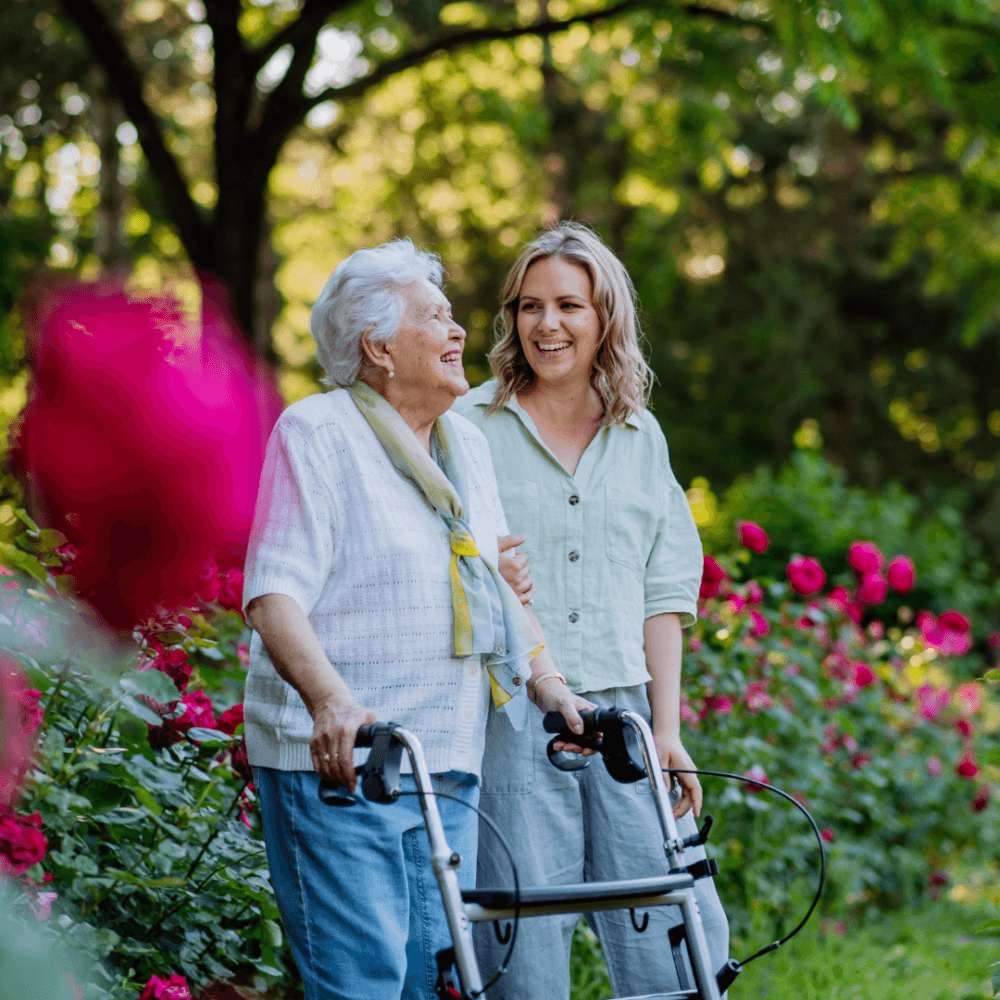 An elderly woman with a walker and a younger woman smile together in a garden filled with pink flowers. - Home Instead