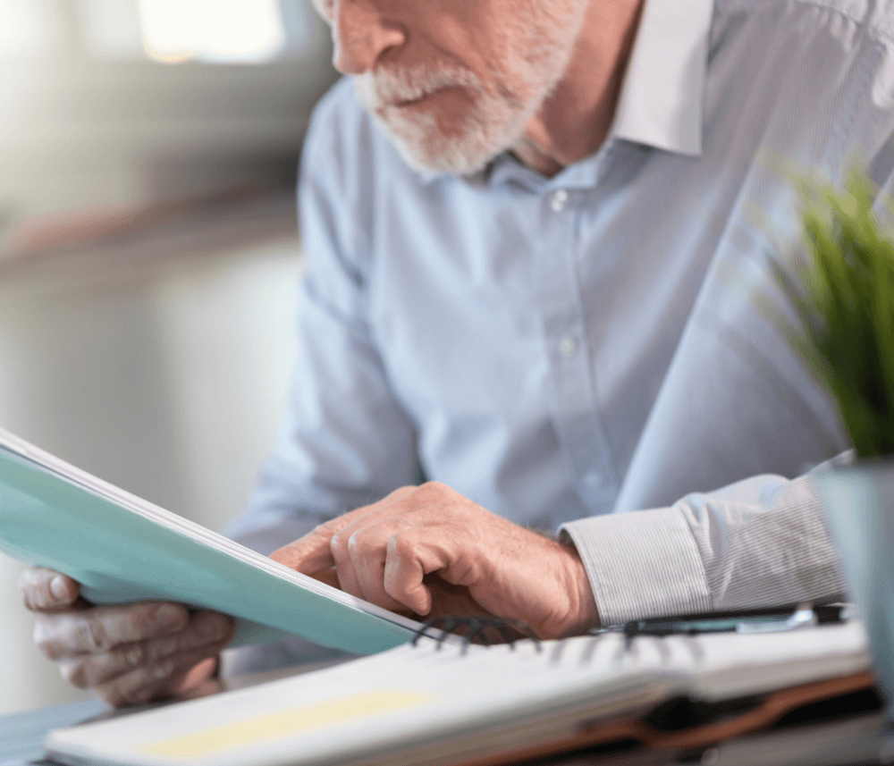 An elderly man in a light blue shirt reviews a document at a desk, with a plant and a notebook nearby. - Home Instead