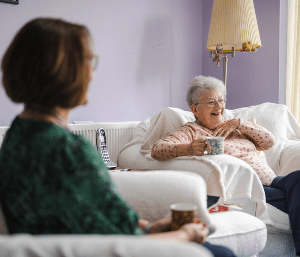 Two elderly women sit and chat in a cozy living room, each holding a mug, with the background softly lit by a floor lamp. - Home Instead