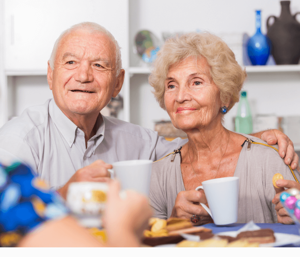 Elderly couple enjoying tea and snacks together, smiling, with other people around a table in a cozy setting. - Home Instead
