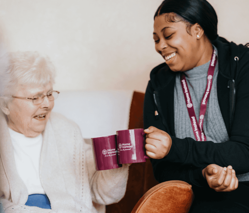 A caregiver and an elderly woman smile and clink purple mugs with 'Home Instead' logos while sitting together on a couch. - Home Instead