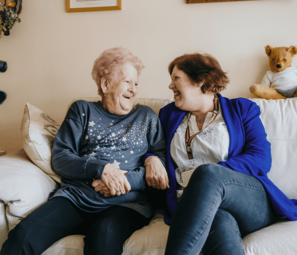 Two elderly women sitting on a couch, holding hands, and laughing joyfully with a stuffed bear in the background. - Home Instead