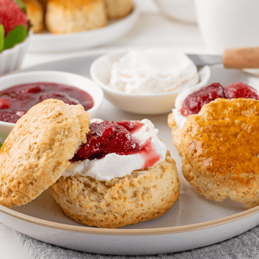 A plate with scones topped with whipped cream and strawberry jam, alongside bowls of extra cream and jam. - Home Instead