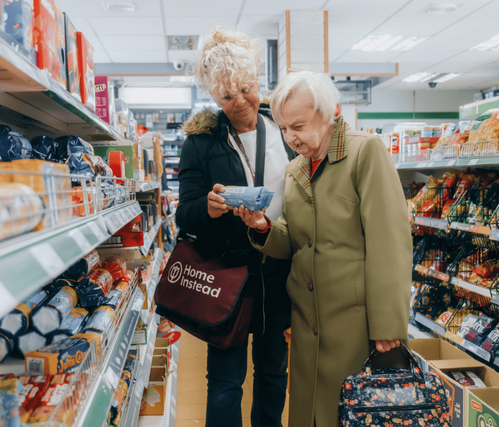 Two women in a store aisle, one holding a package. One carries a "Home Instead" bag. - Home Instead