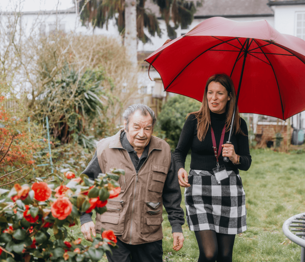 A woman holds a red umbrella while walking with an elderly man in a garden. Red flowers are in the foreground. - Home Instead